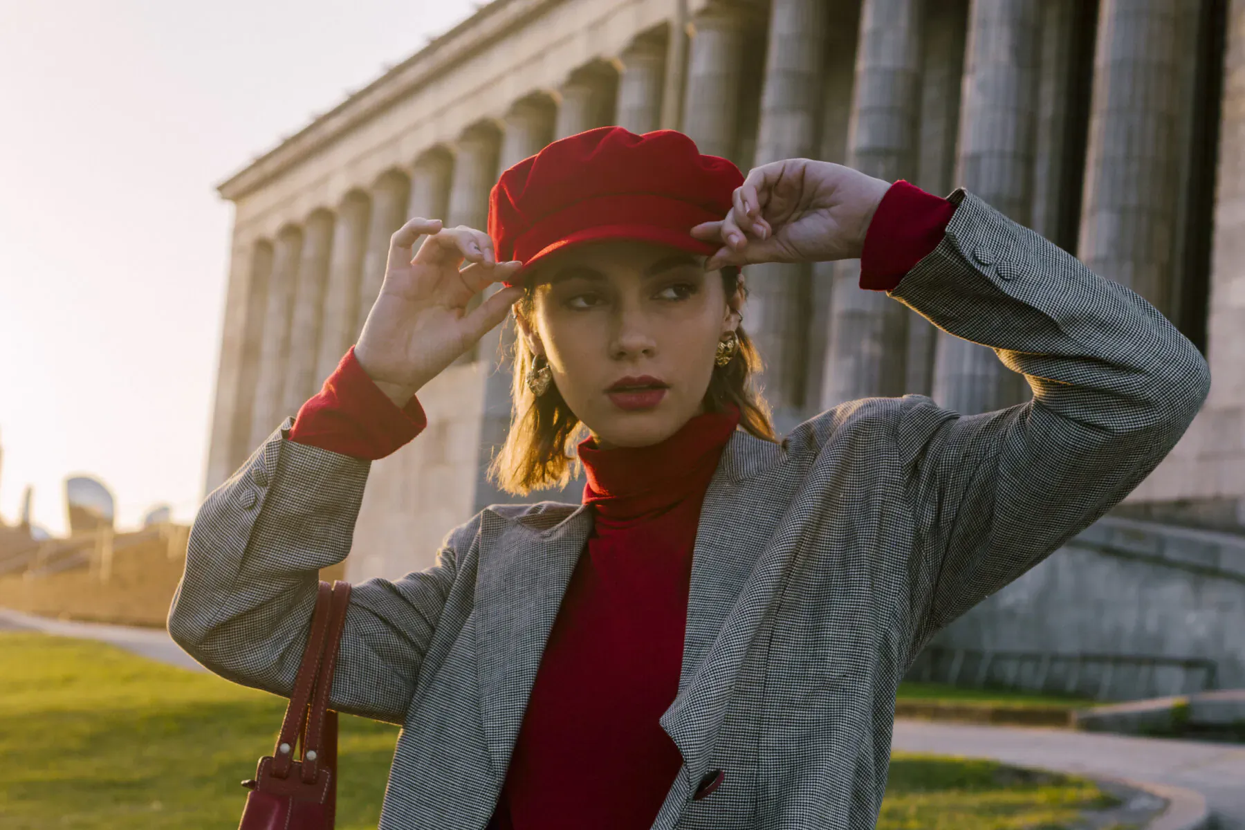 Woman in red beret, red turtleneck, gray blazer and bag adjusts hat in front of grand columns at sunset.