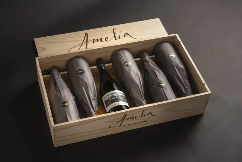 Open wooden box with six 'Amelia' wine bottles wrapped in gray tissue paper, one visible center bottle.