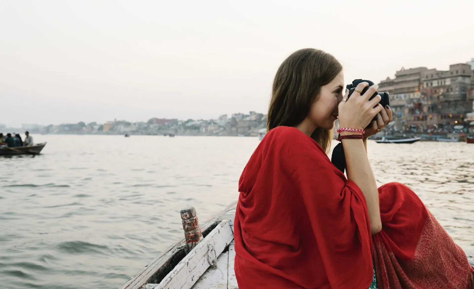 Woman in red shawl photographing with camera from wooden boat on Ganges River, Varanasi ghats in background