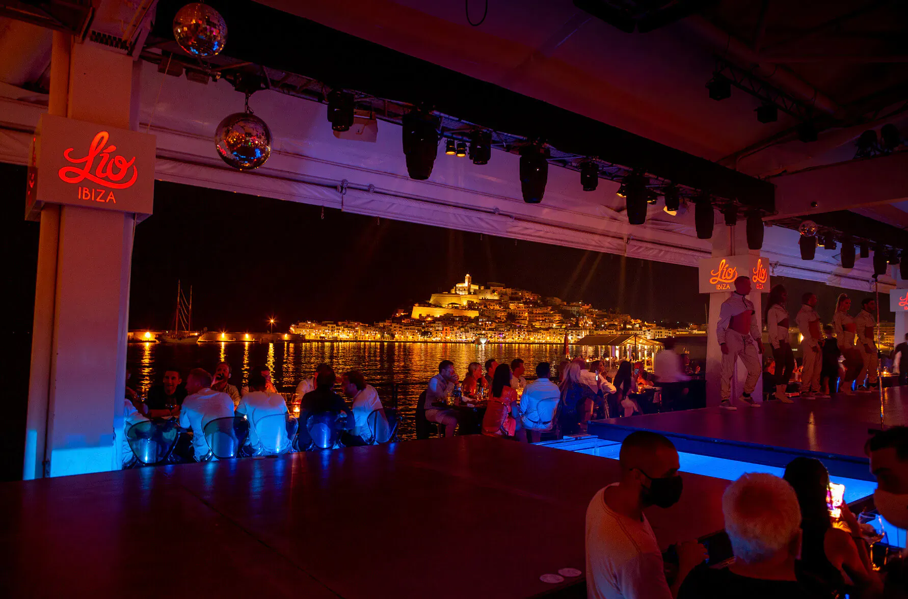 Lío Cabaret Ibiza: neon 'Lio' sign, disco balls, patrons at tables by blue-lit stage, night view of lit-up waterfront palace.