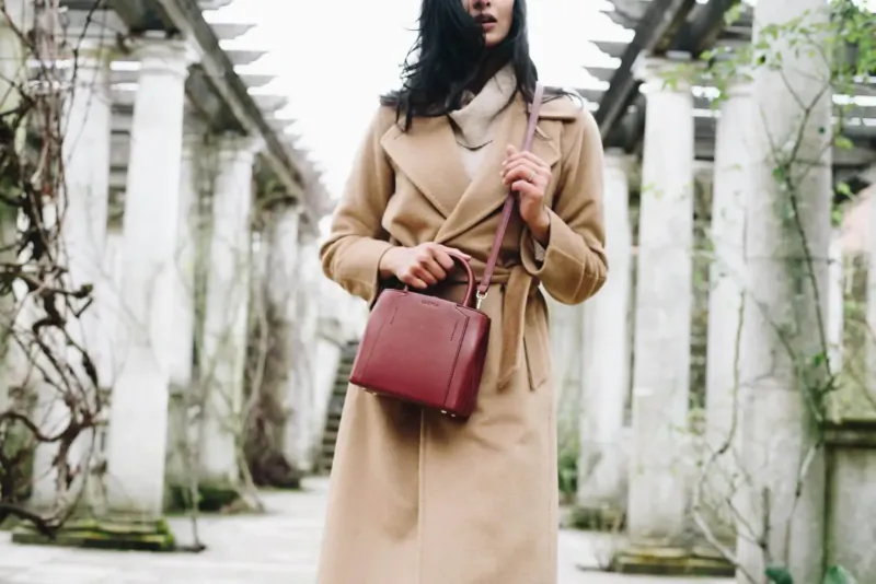 Woman in beige trench coat and scarf holds red leather handbag, standing in vine-covered colonnade