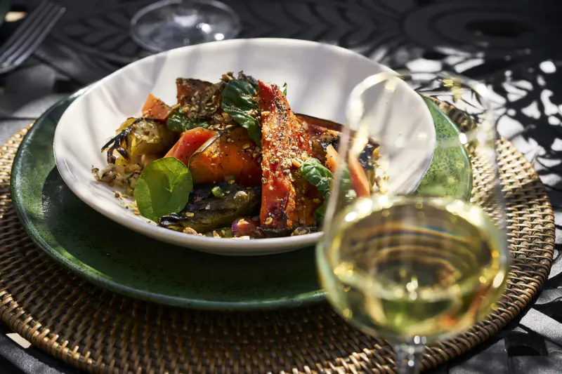 White bowl of roasted vegetables with greens on green plate, white wine glass, outdoor table setting