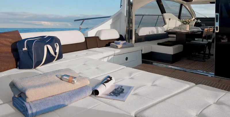 Luxurious yacht deck with white cushions, navy bag, towels, magazine, bottle, and sea view.