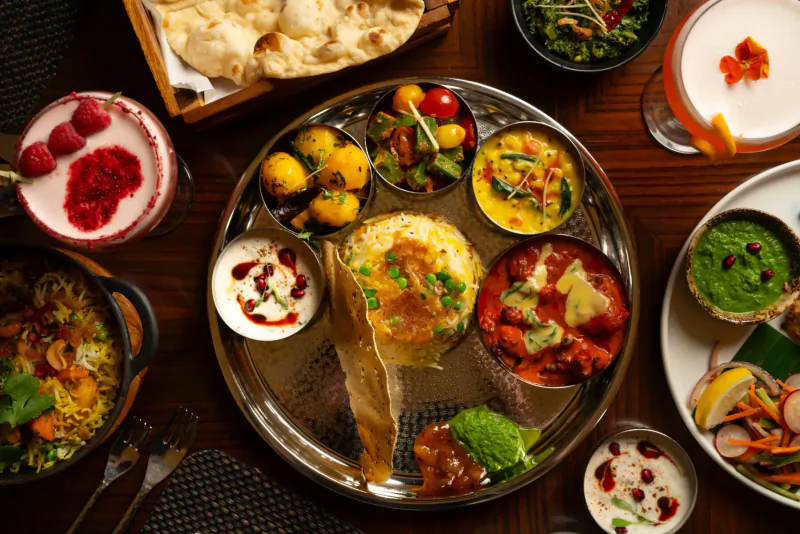 Vibrant Indian thali on steel plate with curries, rice, naan, veggies, and drinks on wooden table