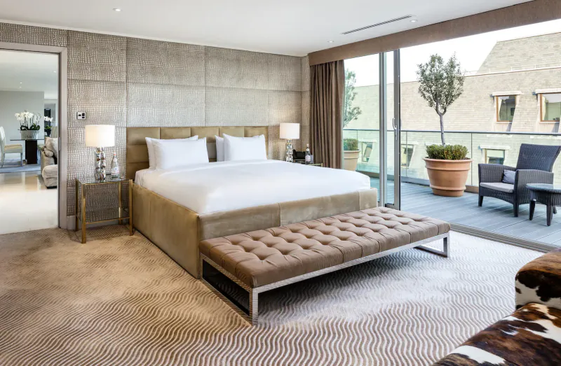 Luxurious hotel bedroom with king bed, beige walls, floor-to-ceiling windows to terrace with potted olive tree and chairs.