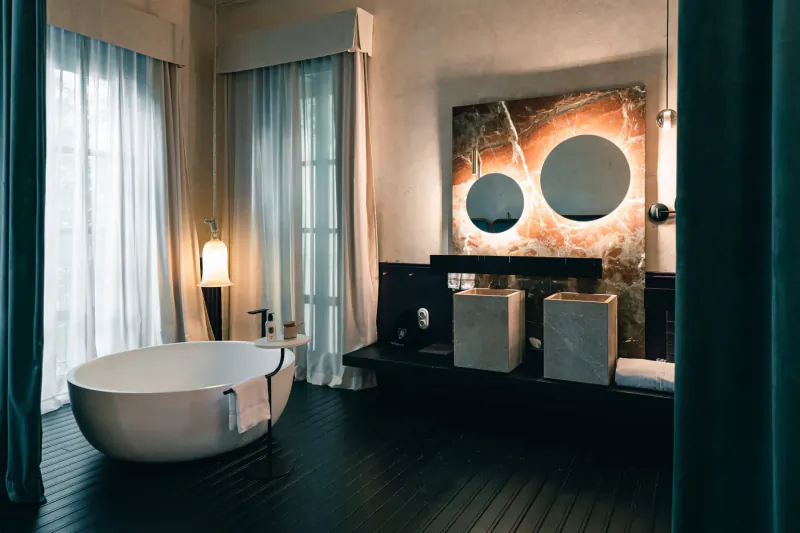 Luxurious bathroom in Can Bordoy Grand House with white freestanding tub, double sinks, round mirrors, and teal curtains