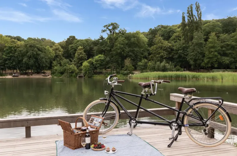 Tandem bicycle with picnic basket of wine and fruit on a wooden pier by lake in lush park, Les Étangs de Corot Paris.