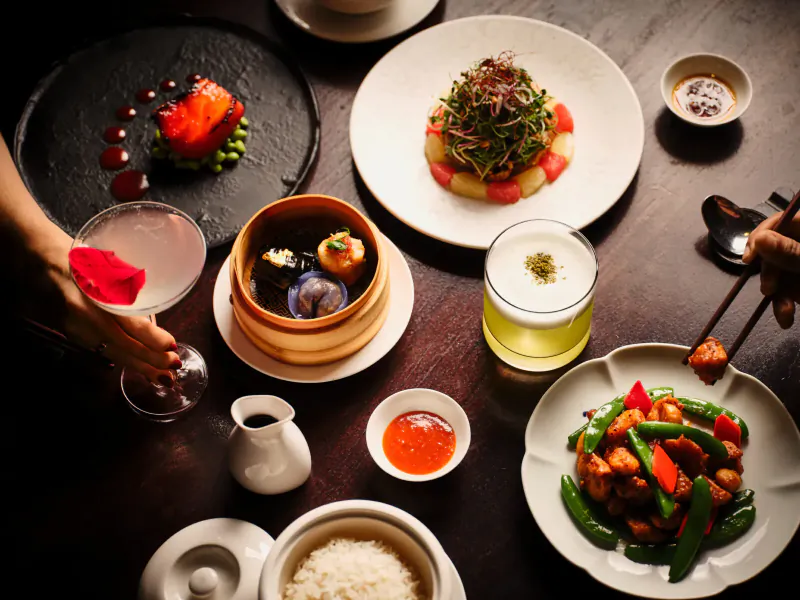Overhead view of Hakkasan Mayfair modern Cantonese dishes: dim sum, kung pao chicken, rice, sauces, cocktail on dark table.