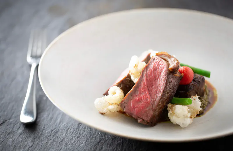 Medium-rare sliced steak with cauliflower puree, green beans, and sauce on white plate beside fork