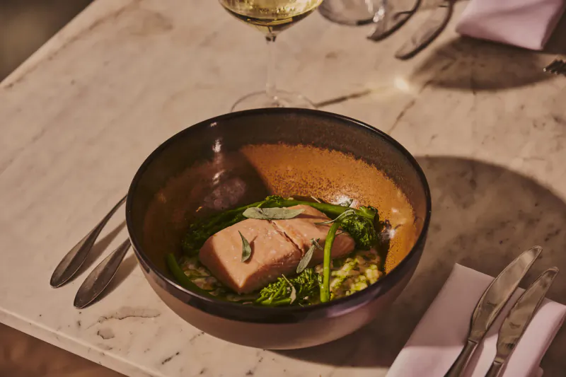 Bowl of poached salmon with greens on mash, silverware, white wine glass on marble table at Treadwell Restaurant
