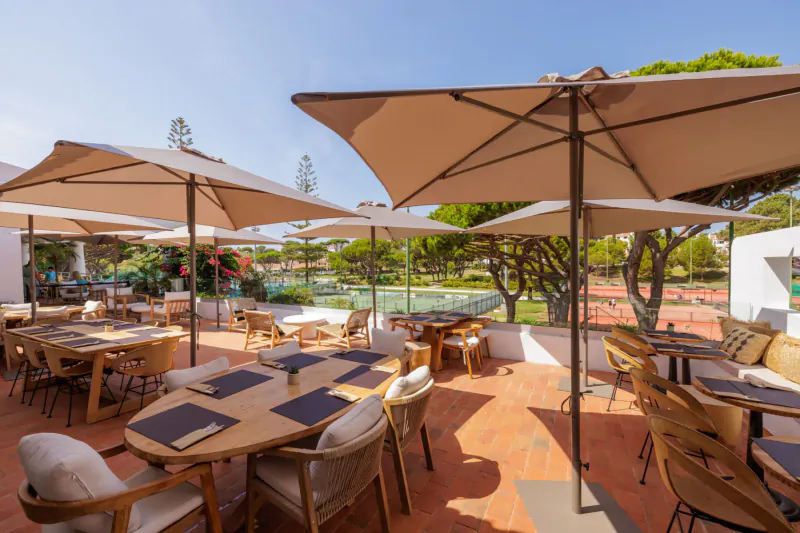 Sunny outdoor terrace restaurant at Vale do Lobo resort with wooden tables, beige umbrellas, and ocean-view pool.