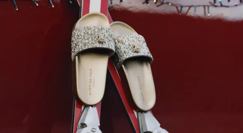 Pair of glittery Kurt Geiger beaded flip-flops leaning against red skis with holiday lights backdrop