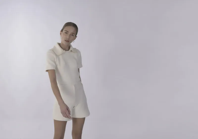 Model poses in white collar short-sleeve romper, luxury plant-based fashion on plain background