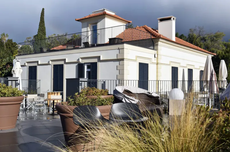 White Les Suites Hotel at The Cliff Bay in Funchal, Madeira, with red-tiled roof, terrace, lounge chairs, umbrellas, and plants under cloudy sky.