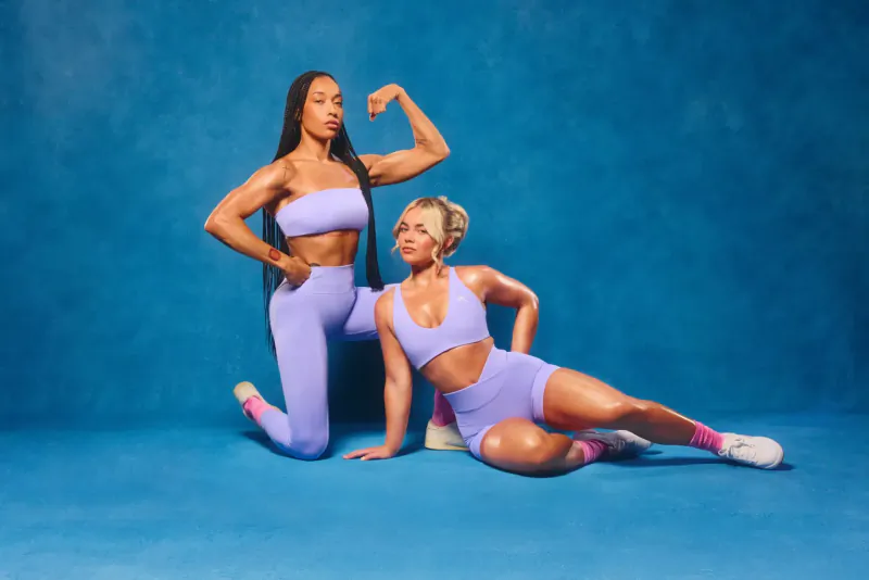 Two women in matching lilac Oner Active outfits flexing muscles on blue background, one standing powerfully, one seated with leg extended.