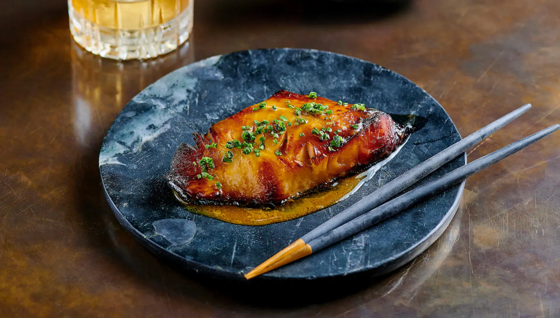 Glazed grilled fish fillet topped with green herbs on dark plate with chopsticks and whiskey glass