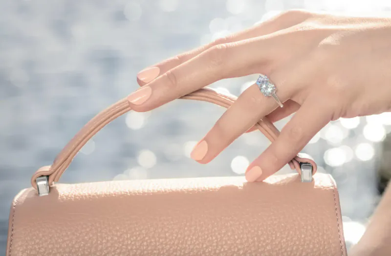 Close-up of woman's hand with peach manicure holding tan leather handbag handle, wearing sparkling diamond ring, Lake Como-inspired backdrop