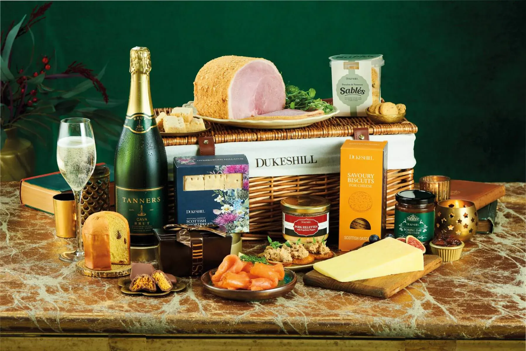 Dukeshill Christmas hamper in wicker basket on green table: champagne, ham, cheese, chutneys, biscuits, and festive foods.
