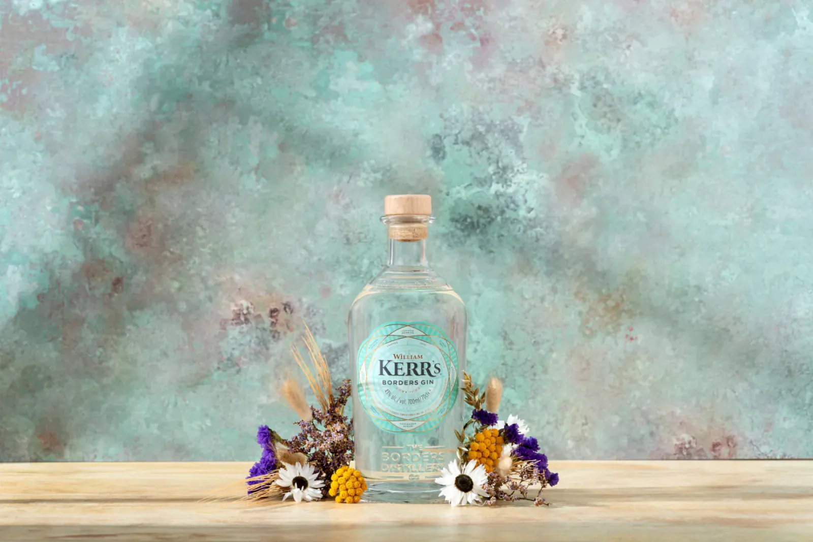 Kerr’s Gin bottle, clear spirit with cork, on wood table amid purple and yellow dried flowers, teal background.