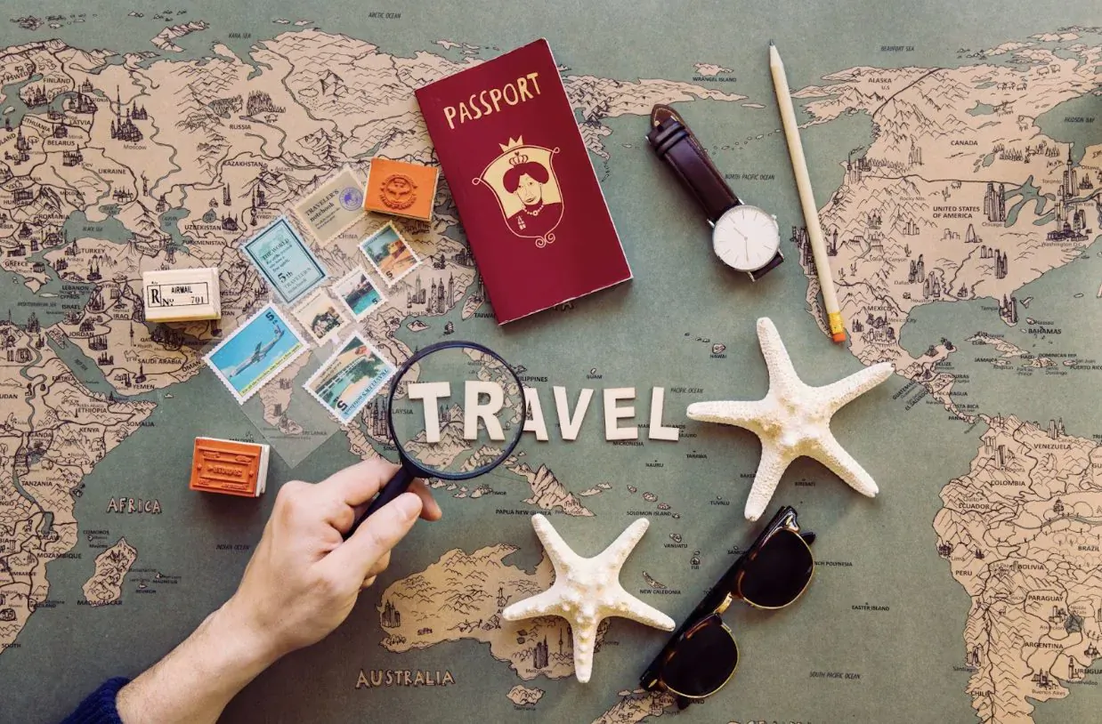 Hand holding magnifying glass over 'TRAVEL' on world map with passport, stamps, seashells, watch, and sunglasses