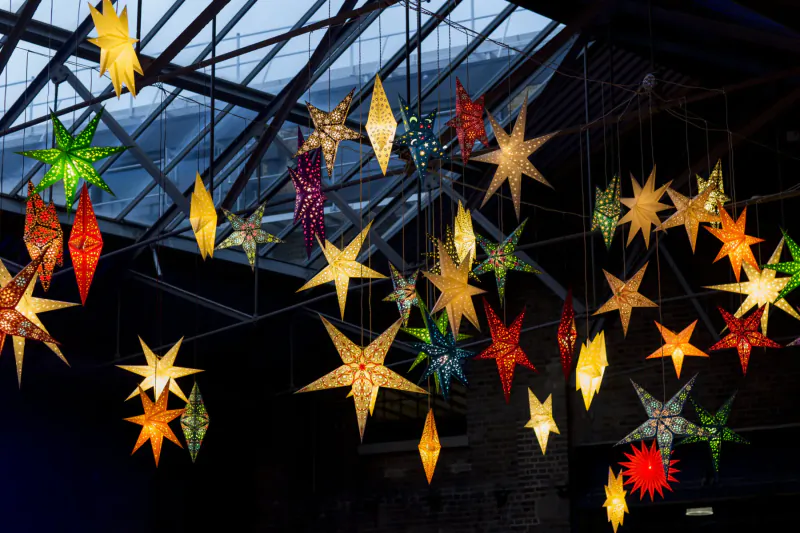 Colorful star-shaped lanterns in various hues hanging from greenhouse ceiling in dim warehouse.