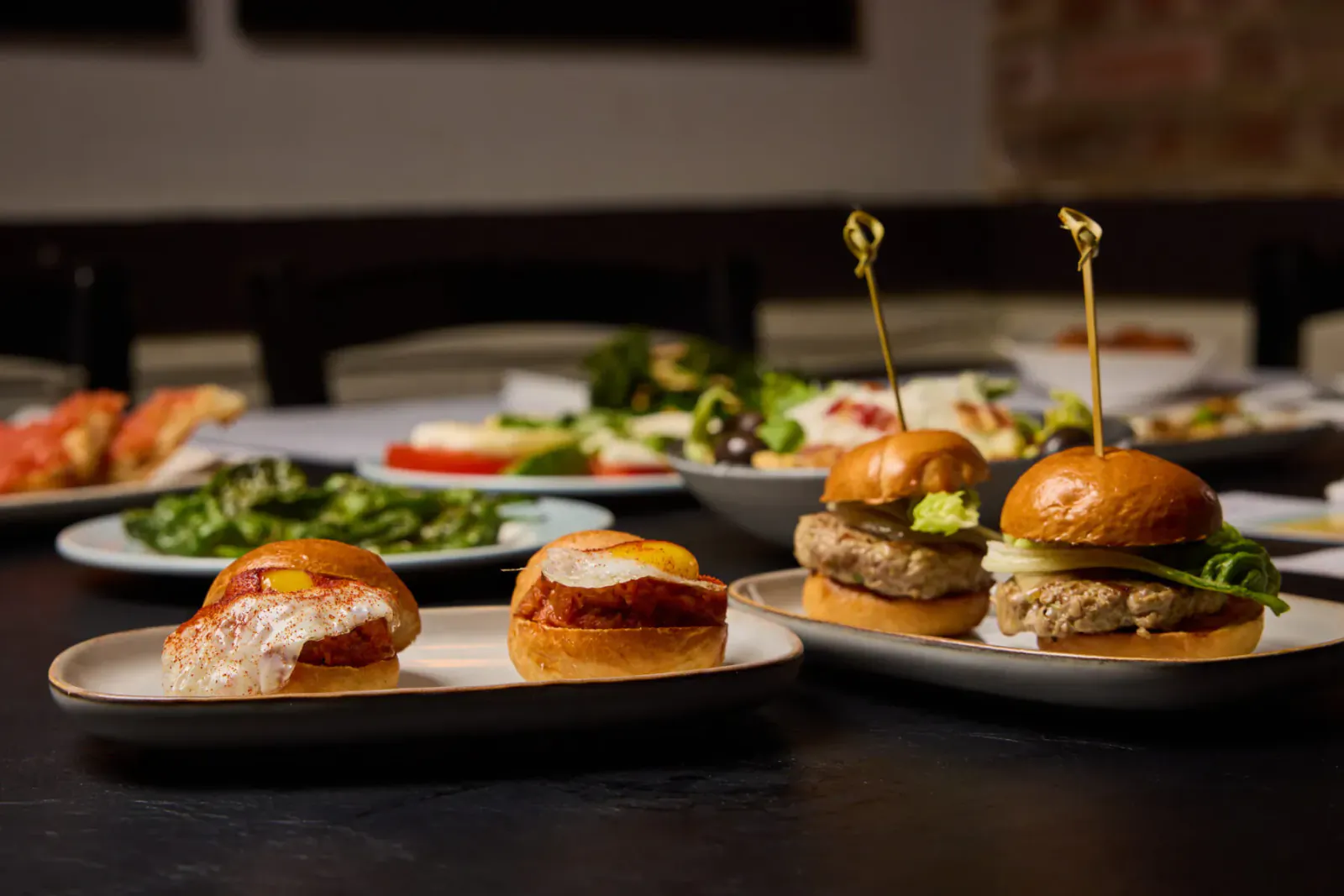 Assortment of mini sliders with beef and tomato toppings on plates, amid Spanish tapas like pizza and salads at El Pirata.