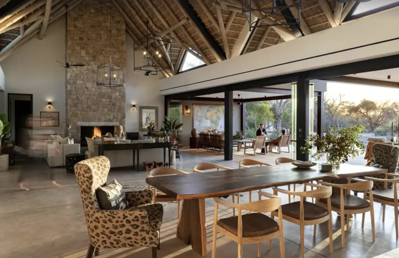 Spacious open-plan lounge and dining area at Monwana Game Lodge with thatched ceilings, stone fireplace, wooden table, leather chairs, and outdoor views.