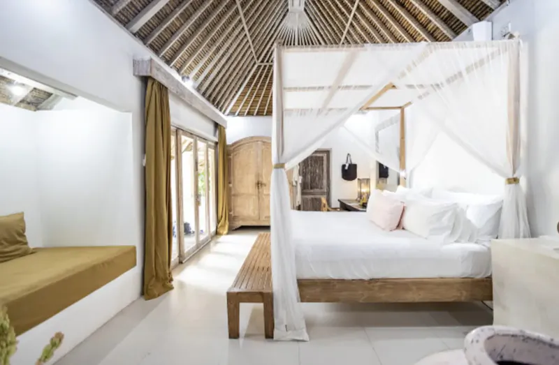 Spacious Bali villa bedroom with thatched ceiling, white canopy bed, gold curtains, and sliding doors to jungle.