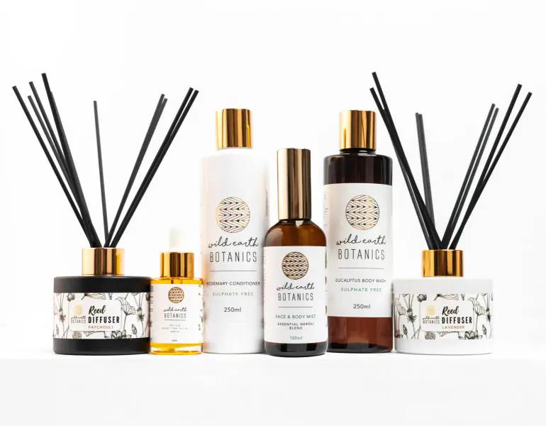 Wild Earth Botanics products: reed diffusers, oils, lotion, serum on white surface with incense sticks.