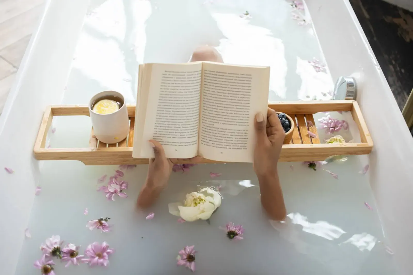 Person relaxing in bathtub with petals, reading open book on wooden bath tray with tea cup