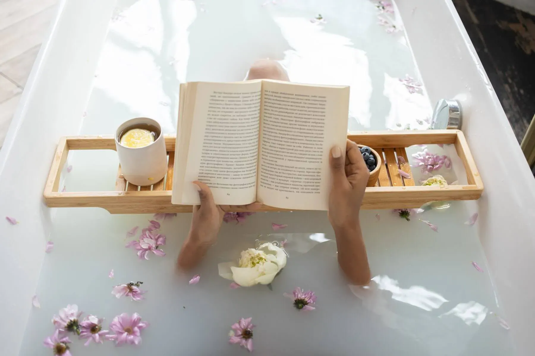 Person relaxing in bathtub with petals, reading open book on wooden bath tray with tea cup