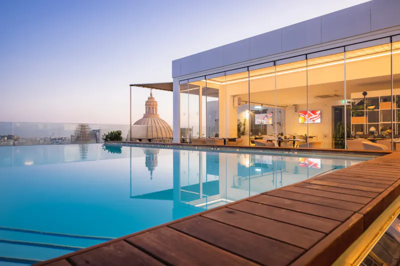 Twilight view of Valletta's rooftop pool and lounge with modern glass building, Buddha statue, and city skyline.