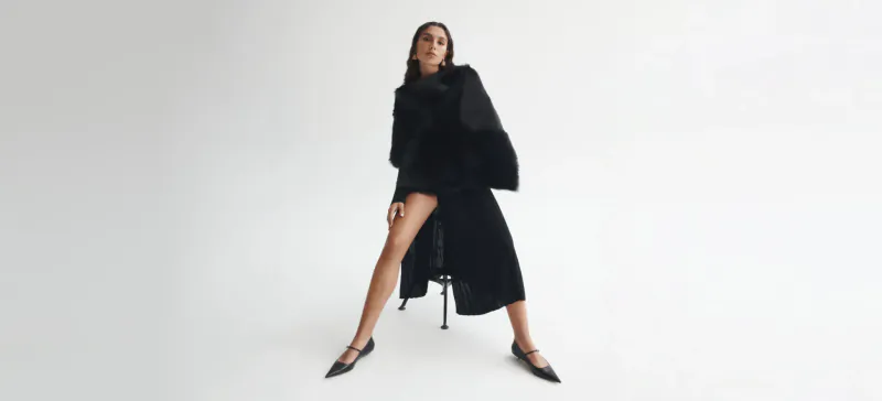 Model in black fur-trimmed coat and heels, posing with legs apart on stool, Scanlan Theodore fashion