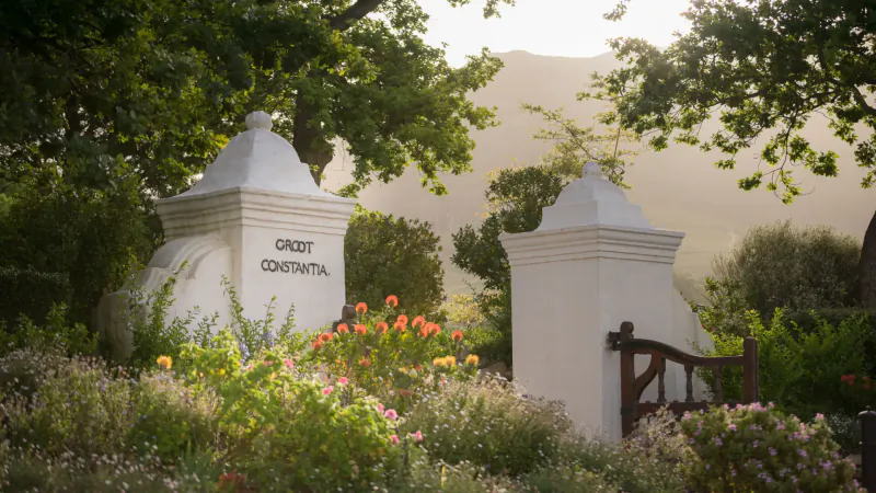 Groot Constantia estate entrance gate with 'Groot Constantia' on white pillars, surrounded by flowers, trees, and mountains.