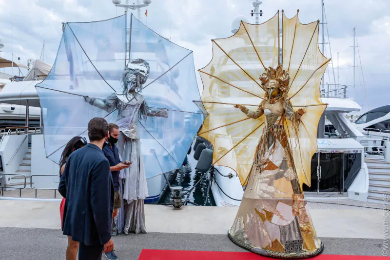 Silver and gold humanoid stilts performers with umbrella frames beside luxury yachts at Balearic event, onlookers watch.