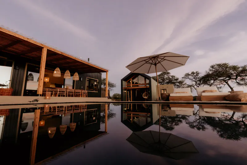 Savanna luxury villas with wooden decks, pendant lamps, umbrella, and reflections in infinity pool at sunset in Namibia wilderness