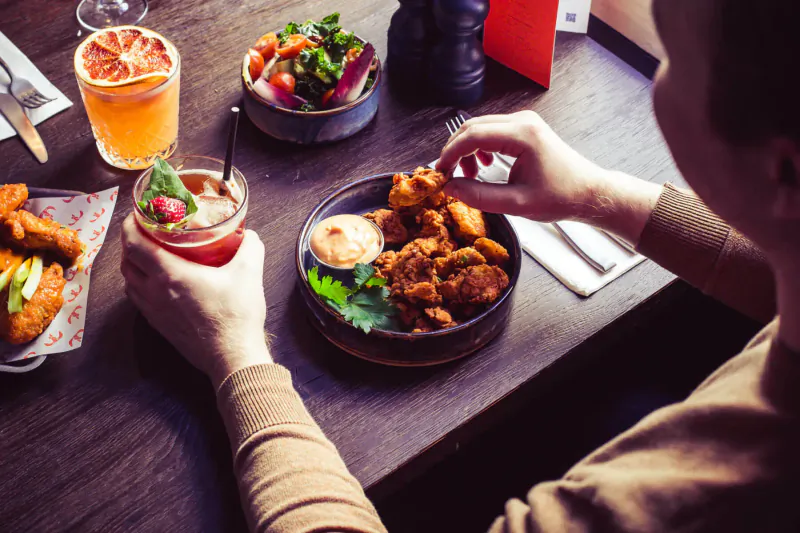 Person eating crispy chicken wings and hummus at CoqBull Soho restaurant table with cocktails and salad