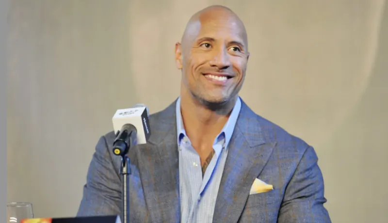 Dwayne 'The Rock' Johnson smiling at podium in gray suit and blue shirt, mic attached.