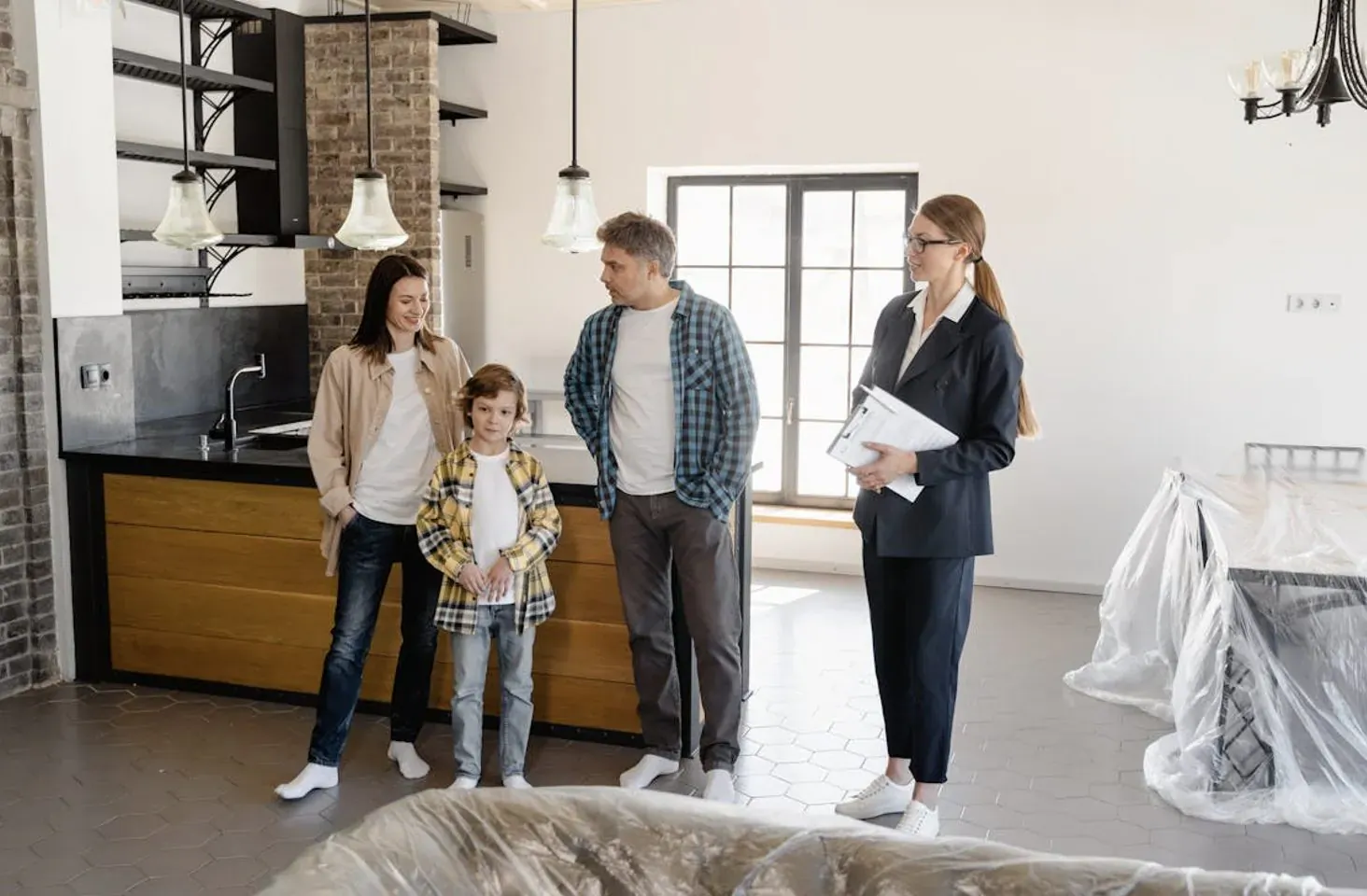 Real estate agent showing luxury kitchen to family: parents and young boy, modern interior with covered furniture