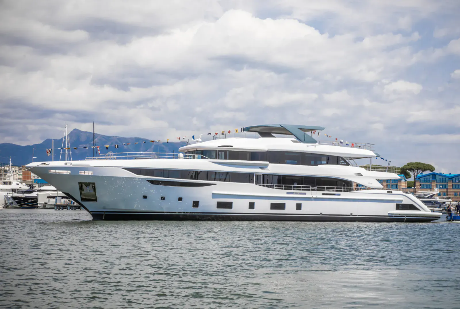 White multi-deck Benetti superyacht docked in marina with mountains and flags, cloudy sky.