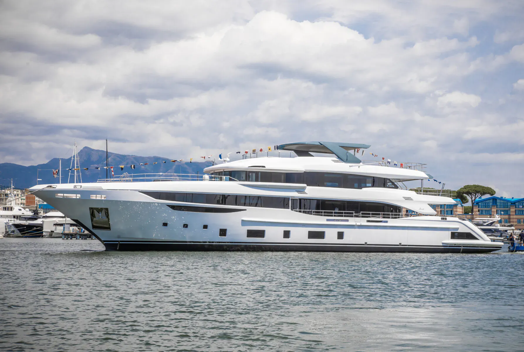White multi-deck Benetti superyacht docked in marina with mountains and flags, cloudy sky.