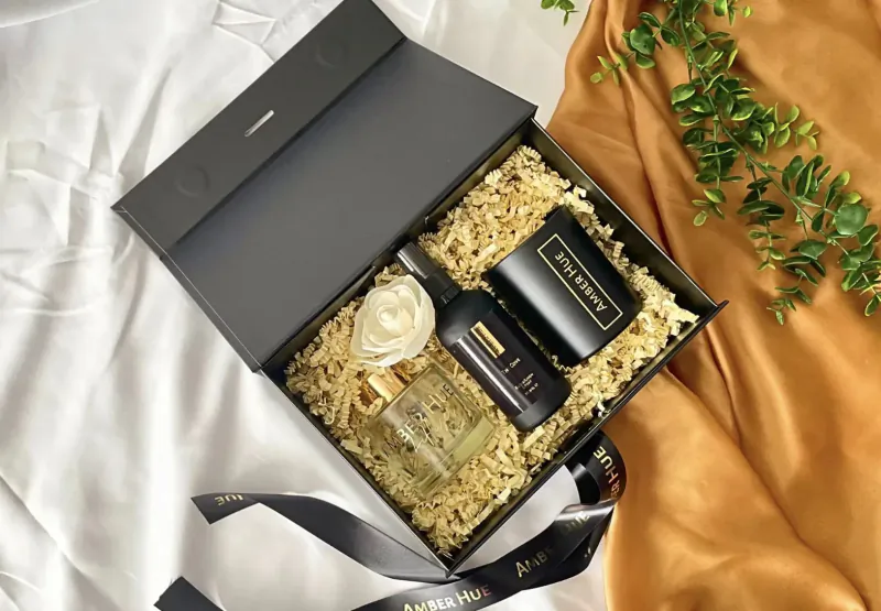 Open black gift box of Amber Hue luxury candles, reed diffuser, and fragrance oil on gold tissue with green leaves.