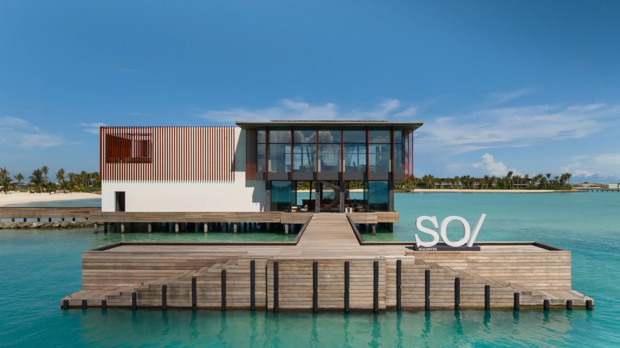 Modern SO/ resort building on wooden platform over turquoise lagoon at Crossroads Maldives, clear blue sky.