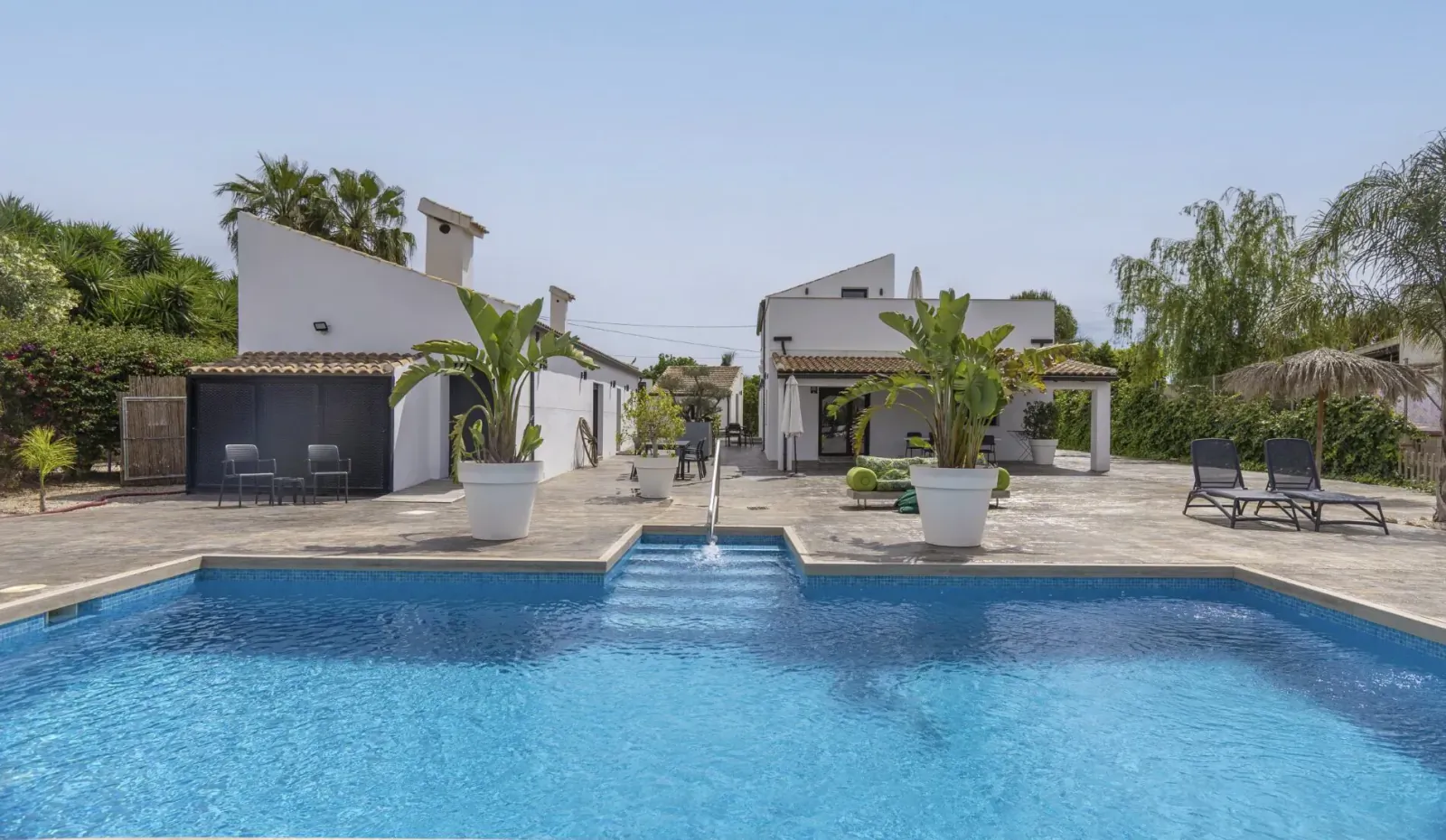 Infinity pool at La Vuelta Boutique Hotel with white villas, palm trees, loungers, and fountain under blue sky.