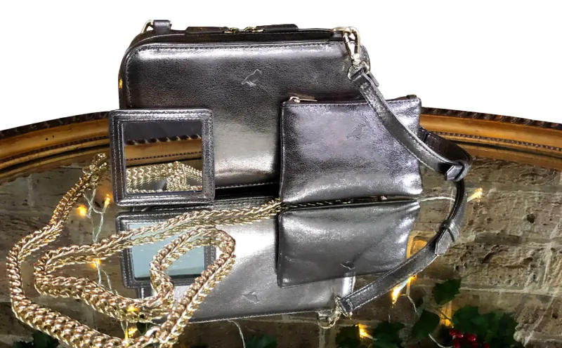 Shiny silver Rufus Country leather handbag, pouch, and mirror on ornate table with gold chain and holiday lights.