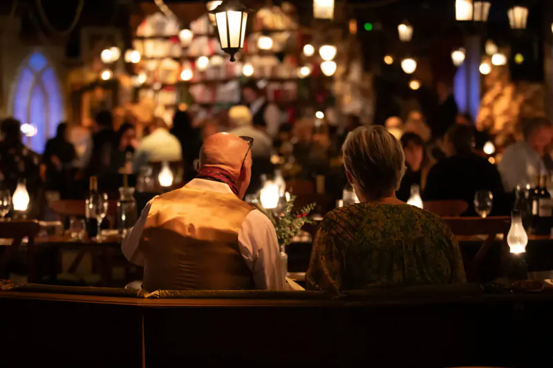 Elderly couple in vest and shawl seated back-to-back at candlelit table in immersive Christmas dining venue with lanterns and crowd.