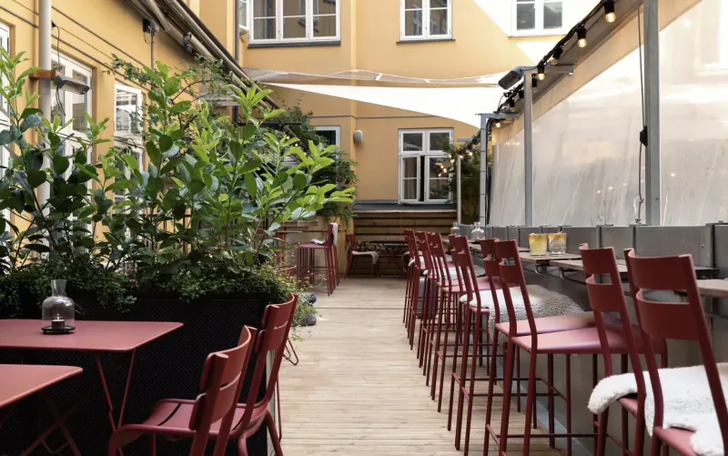 Courtyard at Hotel SP34 in Copenhagen with yellow buildings, lush plants, red chairs, tables, and string lights.