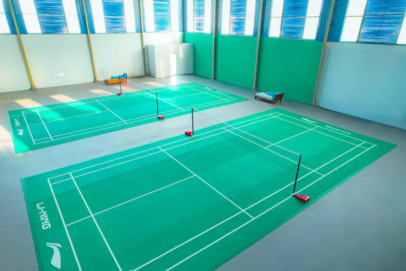 Two indoor badminton courts with green flooring, white nets, and resort gym equipment in the background.
