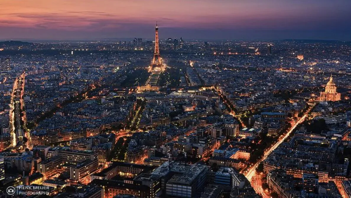 Aerial view of Paris at dusk with Eiffel Tower, lit streets, and city skyline glowing.