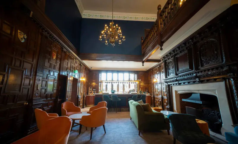 Elegant wood-panelled lounge at Billesley Manor Hotel with chandelier, bar, orange and green armchairs, and fireplace.